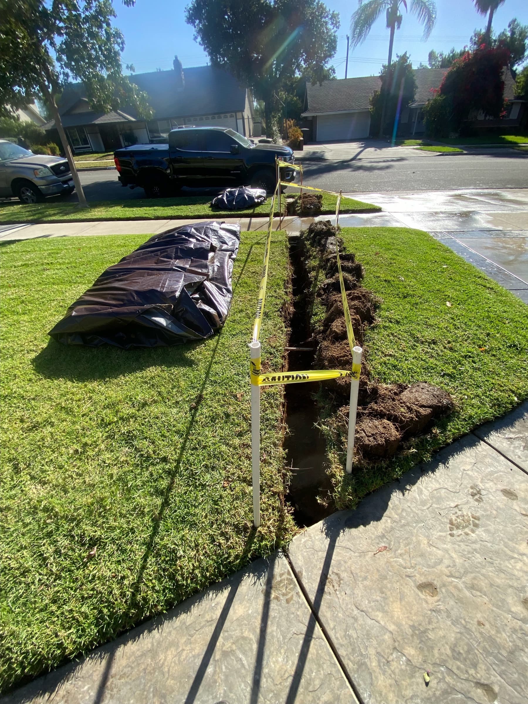 Narrow trench cut through a front lawn from the water meter to the house, with sod set aside and caution tape marking the open line